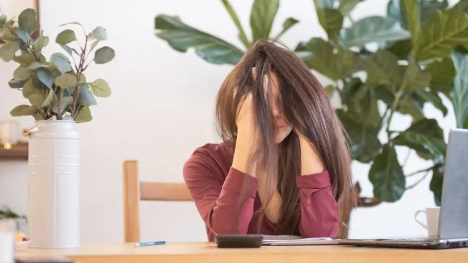 Woman sitting at a desk with her hands over her face looking really stressed. Her hair is a mess and she has work in front of her.